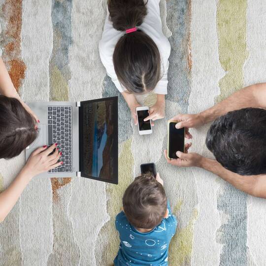 Eine Familie liegt auf dem Teppichboden und jeder schaut auf ein mobiles Endgerät