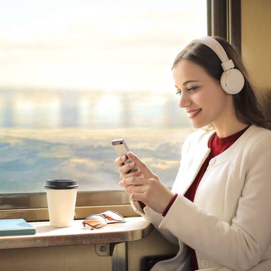 Frau sitzt in Zug mit Kopfhörern auf und schaut auf Smartphone-Display