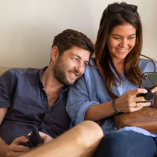 Zwei Personen nutzen Smartphone, junger Mann lehnt sich dabei lächelnd an junge Frau an