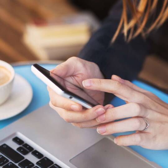 Frau sitzt mit Smartphone in der Hand vor Laptop und Kaffeetasse