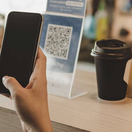Person hält Smartphone in der Hand, dahinter ist ein Barcode, Coffee To Go und eine braune Tüte