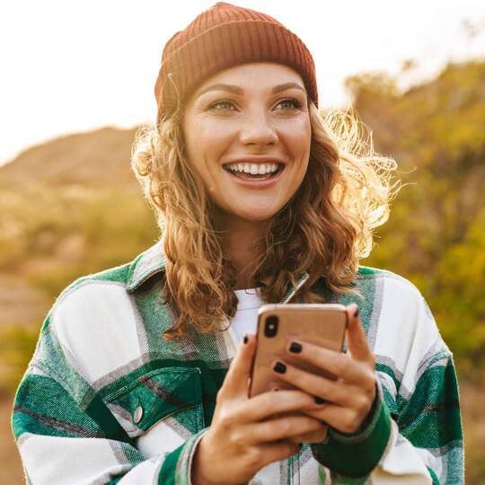 Frau mit Mütze und Hemd lächelt und hält Handy in der Hand