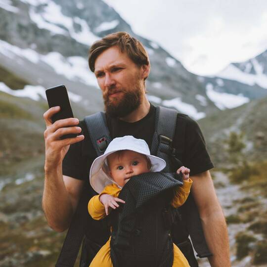 Mann mit Kind im Tragegurt in den Bergen schaut auf sein Smartphone
