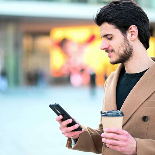 Person steht draußen mit einem Kaffee und einem Smartphone in der Hand