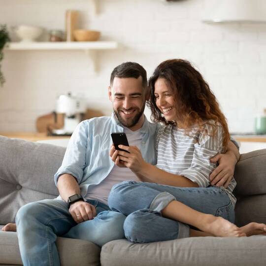 Paar sitzt lächelnd auf Couch und schaut gemeinsam auf ein Smartphone