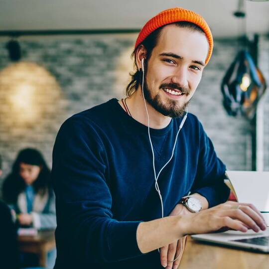 Junger Mann mit Mütze und Kopfhörern sitzt mit Laptop an öffentlichem Ort und lächelt