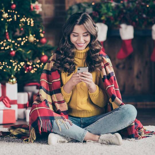 Frau sitzt lächelnd mit einer Decke über den Schultern mit ihrem Smartphone vorm Weihnachtsbaum