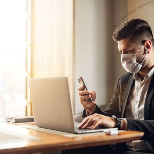 Mann mit Gesichtsmaske sitzt vor Laptop und Smartphone