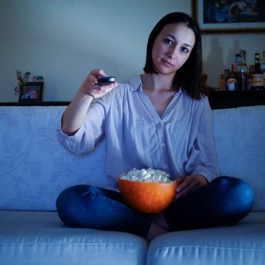 Frau sitzt mit Popcorn auf Couch getaucht in blaues Fernseherlicht