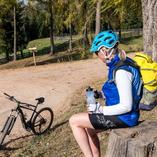 Frau sitzt. Trägt Sportkleidung und einen Fahrradhelm. Im Hintergrund steht ein E-Bike.