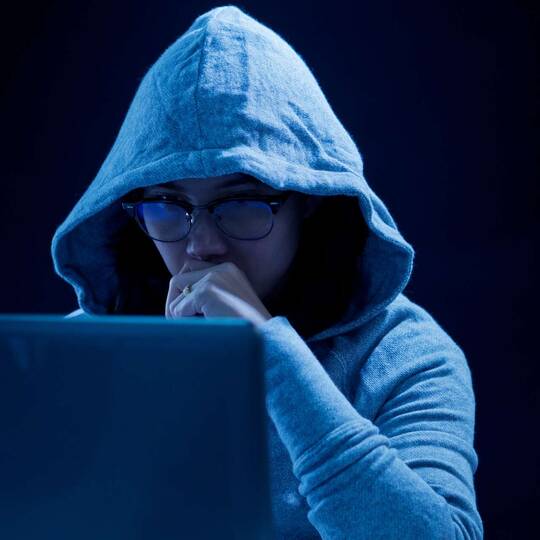 Person mit Kapuze in dunkler Umgebung schaut auf Laptop