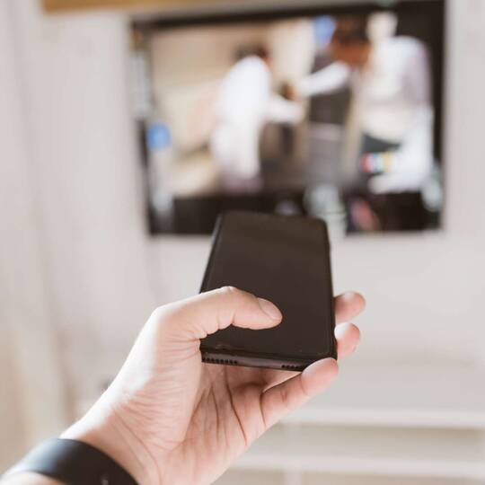 Hand mit Smartphone streckt in Richtung Fernseher