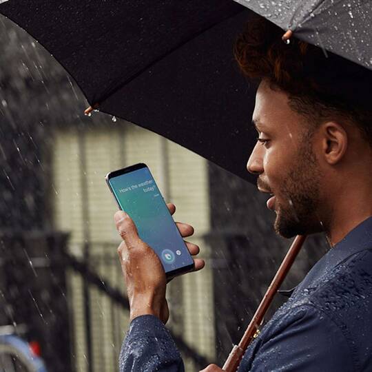 Ein Mann steht im Regen mit einem Regenschirm und hält ein Samsung Smartphone in der Hand, schaut auf das Display um den Sprachassistenten Bixby zu bedienen