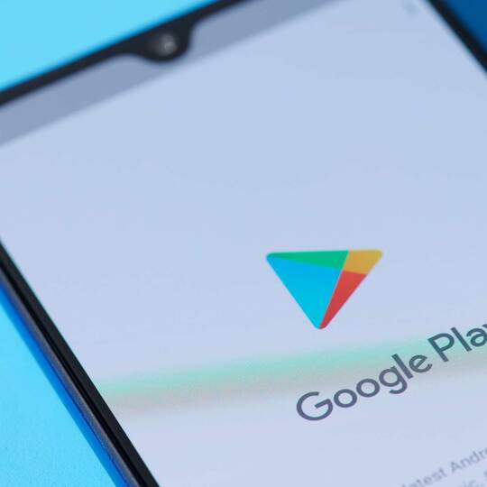 Schwarzes Smartphone mit dem Google Play Startbildschirm auf einem hellblauen Hintergrund.