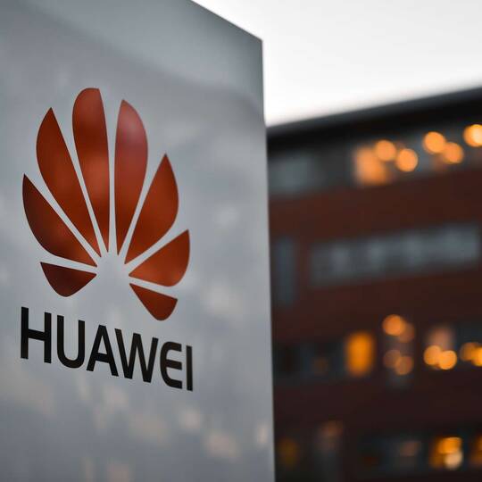 Logoabbild der Firma Huawei, im Hintergrund ein beleuchtetes Gebäude.