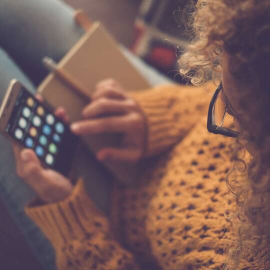 Person im Pulli tippt gemütlich im Sitzen mit zwei Händen auf Smartphone