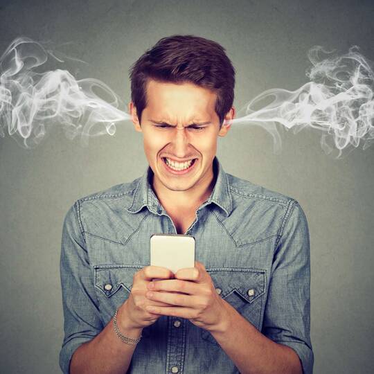 Mann guckt verärgert auf sein Smartphone, während ihm Rauchwölkchen aus den Ohren steigen