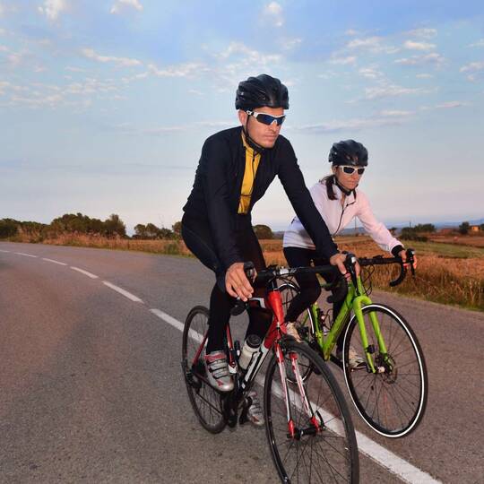 Zwei Radfahrer in Fahrradbekleidung sind auf asphaltierter Straße unterwegs.