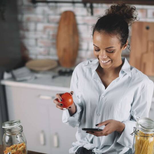 Frau in Küche schaut fröhlich auf Smartphone Displa