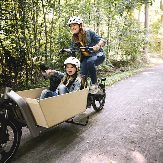 Frau fährt mit Kind und Helm auf Lastenrad durch den Wald
