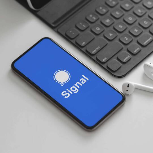 Smartphone mit Signal-Logo auf Display liegt neben AirPods und Tastatur