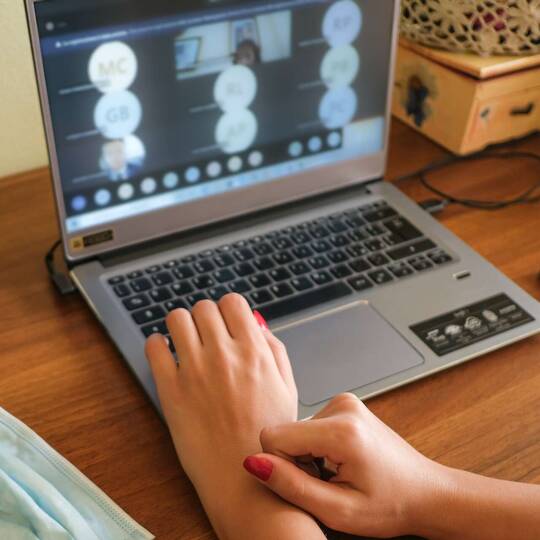 Laptop mit Microsoft Teams Videocall auf Display, Hände liegen davor auf Tisch neben Maske