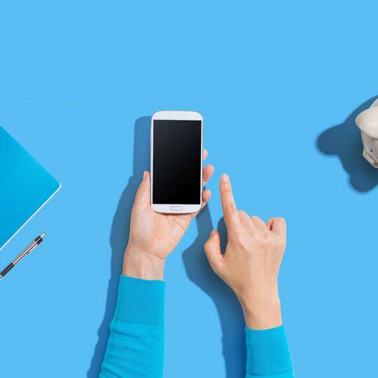 Zwei Hände halten Smartphone vor blauem Hintergrund mit Portemonnaie und Sparschwein