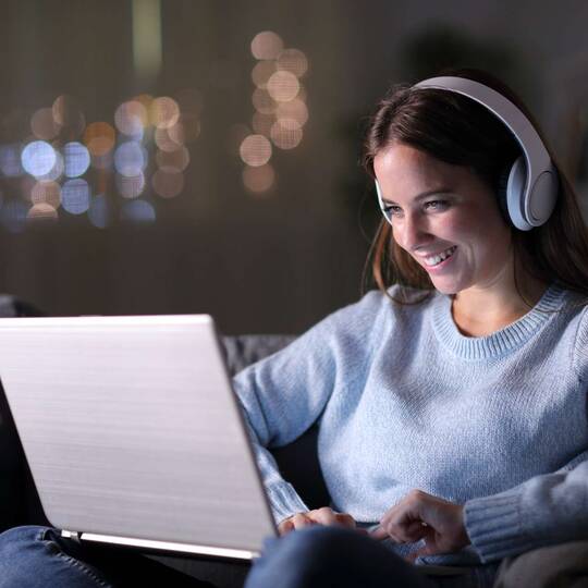 Frau mit großen Kopfhörern sitzt lächelnd vor Laptop in dunkler Umgebung