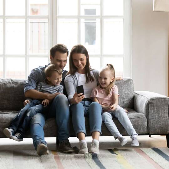 Familie mit zwei Kindern sitzt auf Sofa und schaut auf Smartphone