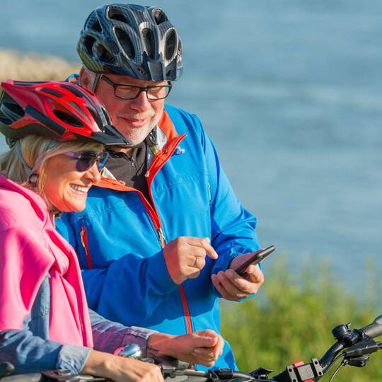 Zwei ältere Menschen am Wasser mit Fahrrädern und Fahrradkleidung schauen auf Handy