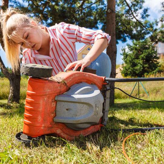 Eine Frau guckt sich die Unterseite eines Rasenmähers an