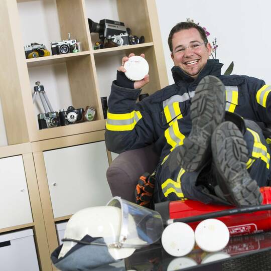 Mann in Feuerwehranzug sitzt lächelnd in Büro, auf Tisch vor ihm liegen Helm und Feuermelder