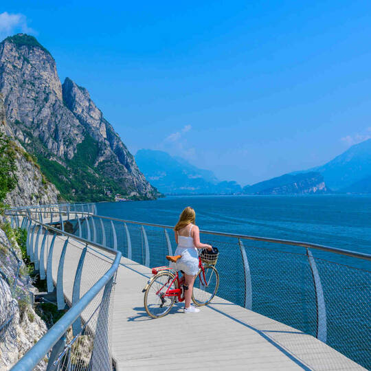 Frau mit Rad steht auf einer Brücke vor See in bergiger Landschaft