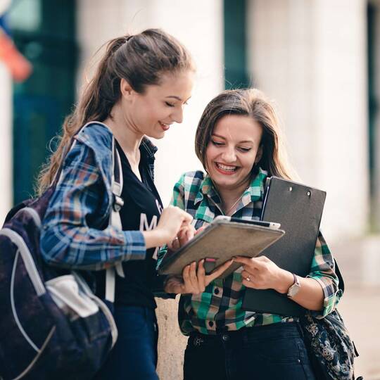 Zwei Schülerinnen stehen mit iPad vor halbhoher Mauer und schauen fröhlich auf das Tablet