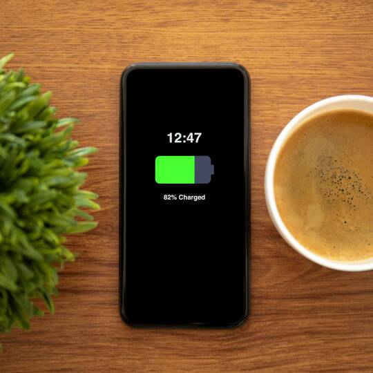 Ladendes Handy liegt ohne Kabel auf Tisch zwischen Plastikpflanze und Kaffeetasse