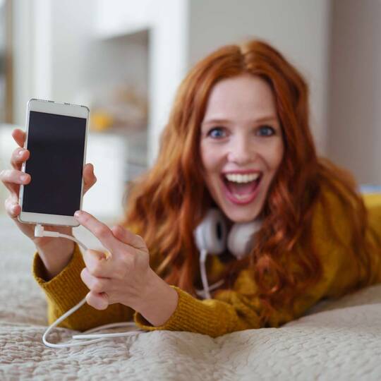 Frau liegt mit Kopfhörern auf einem Bett und zeigt freudig auf ihr Smartphone