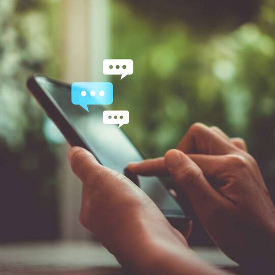 Zwei Hände tippen auf Smartphone mit virtuellen Chat-Textblasen als Hologramm um das Smartphone herum