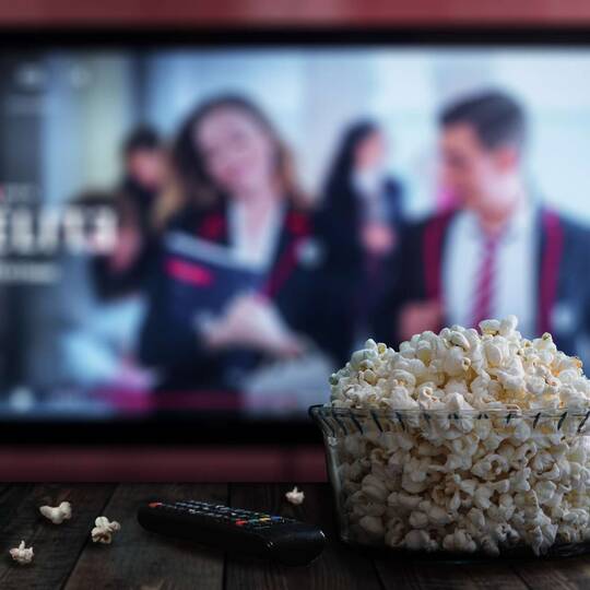 Fernseher mit Netflix im Hintergrund, im Vordergrund sind Popcorn und Fernbedienung