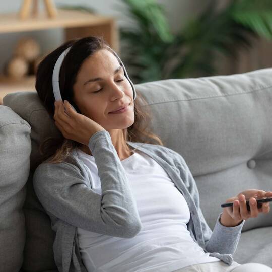 Frau lauscht mit geschlossenen Augen ihren Kopfhörern, welche mit einem Smartphone verbunden sind.