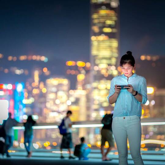 Eine Eine Frau steht vor der Skyline von mehreren Hochhäusern, sie schaut auf ihr Smartphone in ihrer Hand.