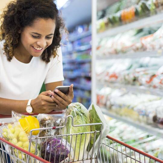 Frau mit Einkaufswagen im Supermarkt schaut auf ihr Handy