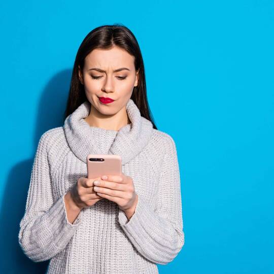 Junge Frau steht vor einem hellblauen Hintergrund und schaut verwundert auf ihr Smartphone in ihrer Hand.