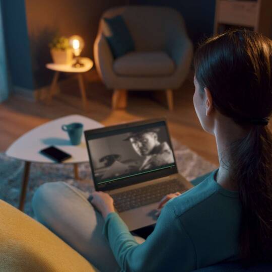 Frau sitzt in Wohnzimmer vor Laptop und schaut dort ein Video