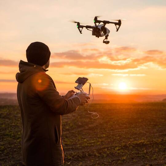 Person steuert Drohne mithilfe einer Fernbedienung auf Feld bei Sonnenuntergang