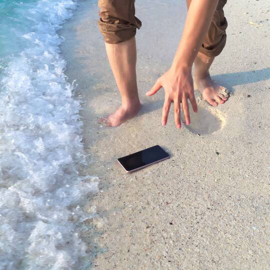 Person greift nach Smartphone im Sand, welches gleich von einer Welle überspült wird