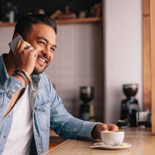 Mann telefoniert freundlich lächeldn im Sitzen mit Kaffee vor sich stehend
